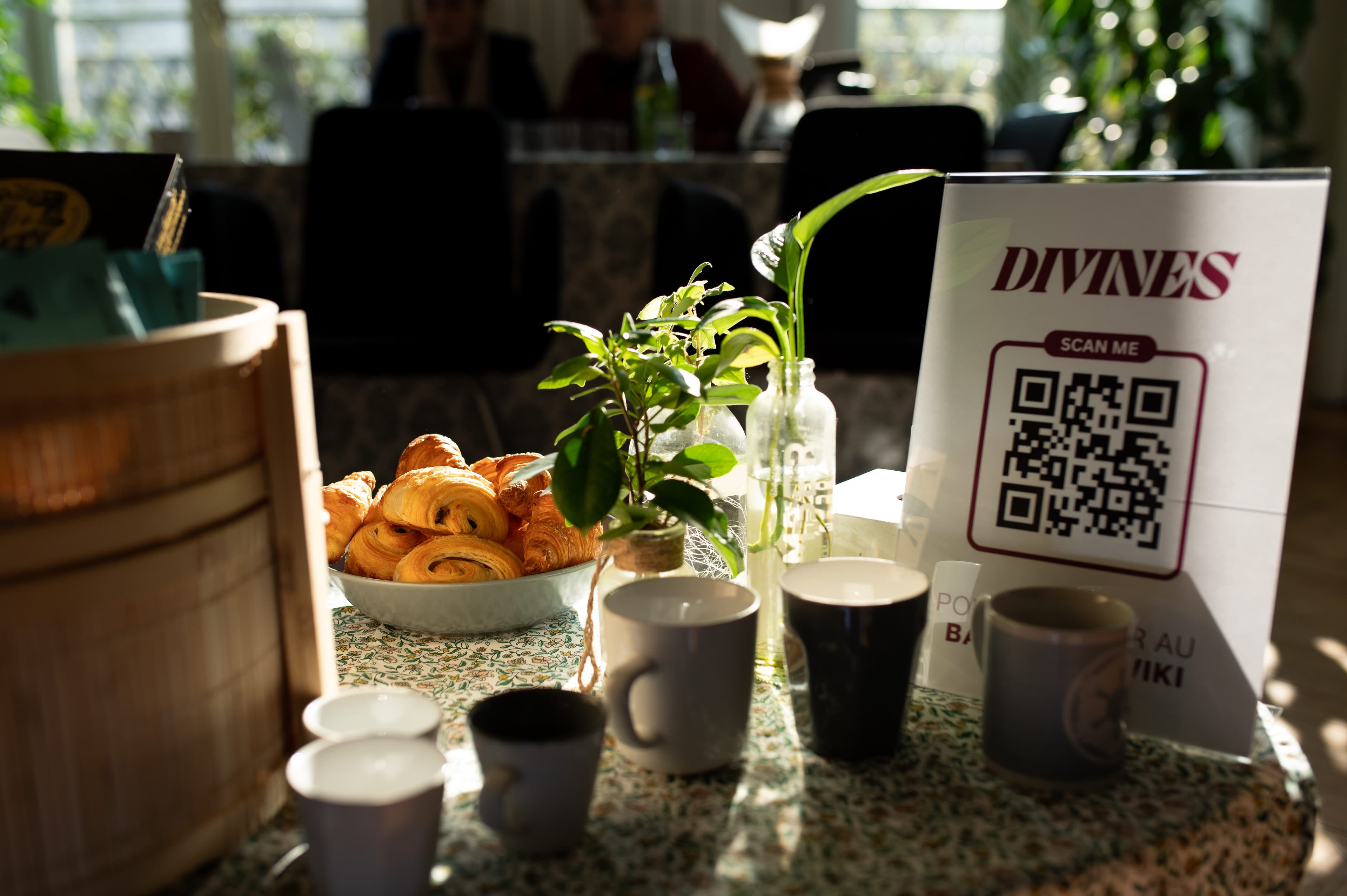 Table d'accueil du Base Camp : panneau Divines avec QR code, viennoiseries, tasses et boutures dans la lumière du matin.