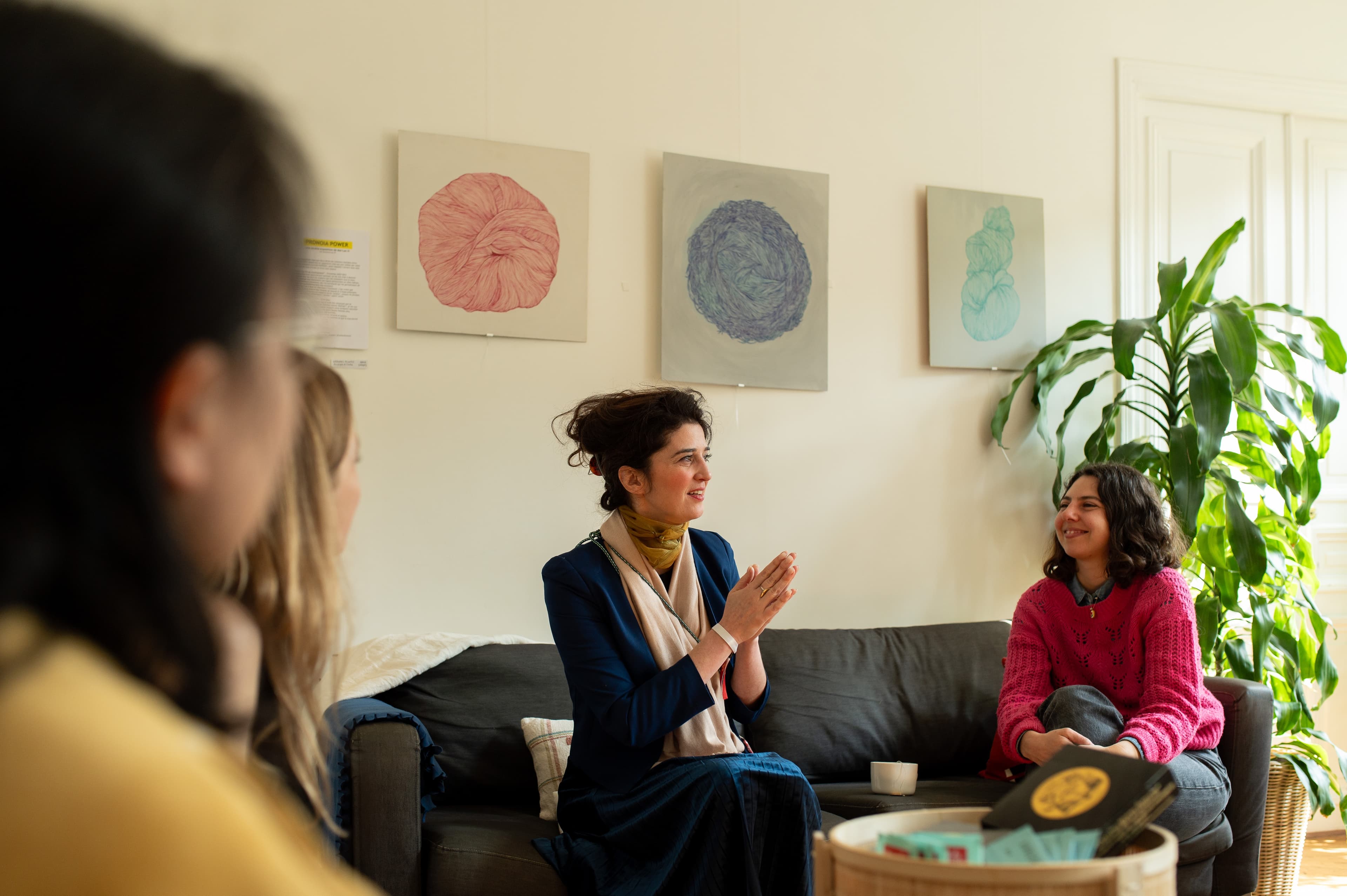 Trois femmes en conversation sur un canapé, devant une série de tableaux abstraits.