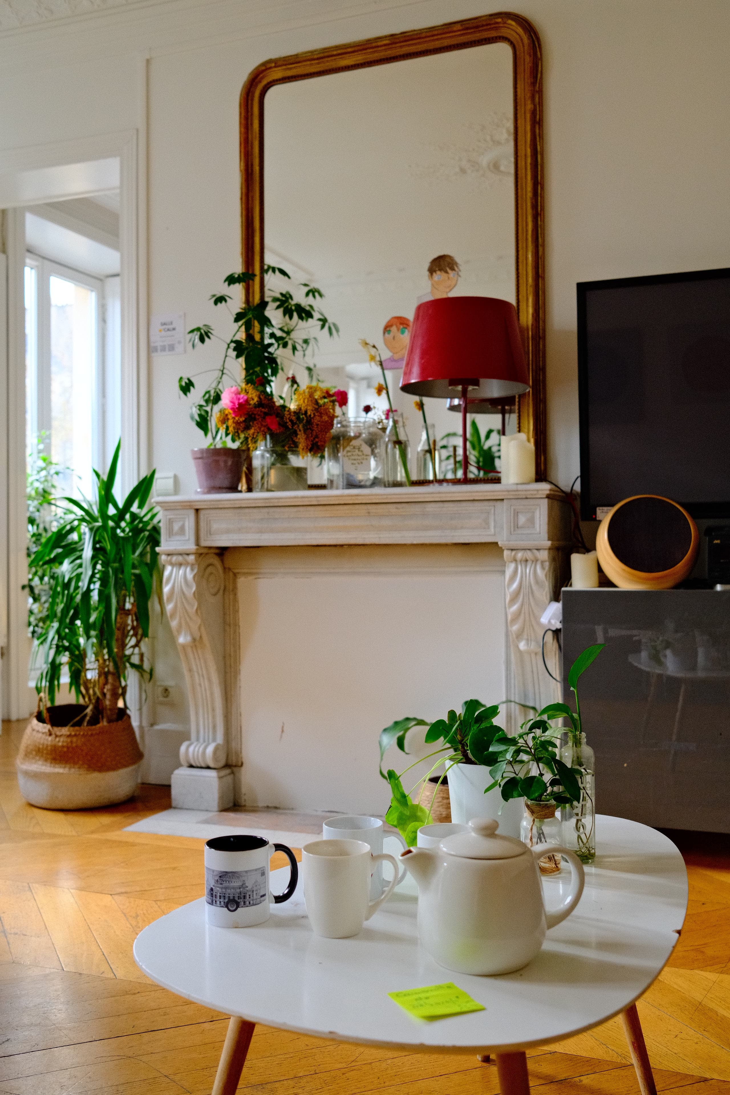 Cheminée en marbre surmontée d’un grand miroir doré, bouquet de fleurs, lampe rouge et plantes dans un salon parisien.