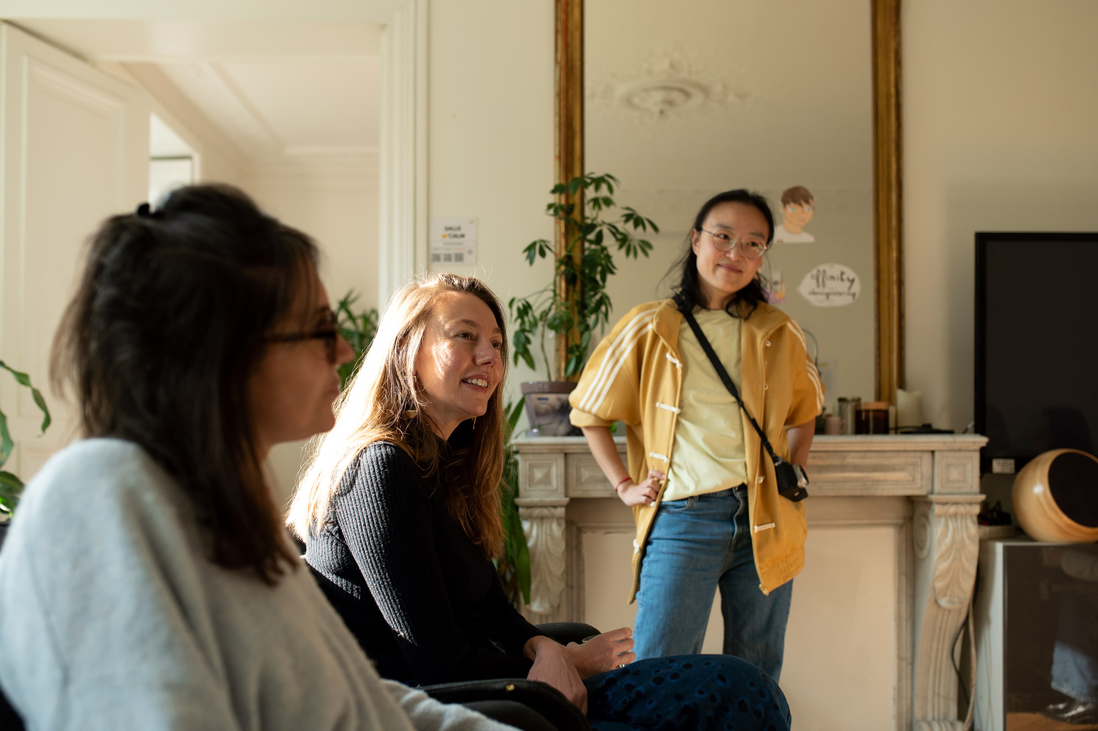 Groupe de femmes en discussion, l’une debout en veste jaune devant un miroir doré.